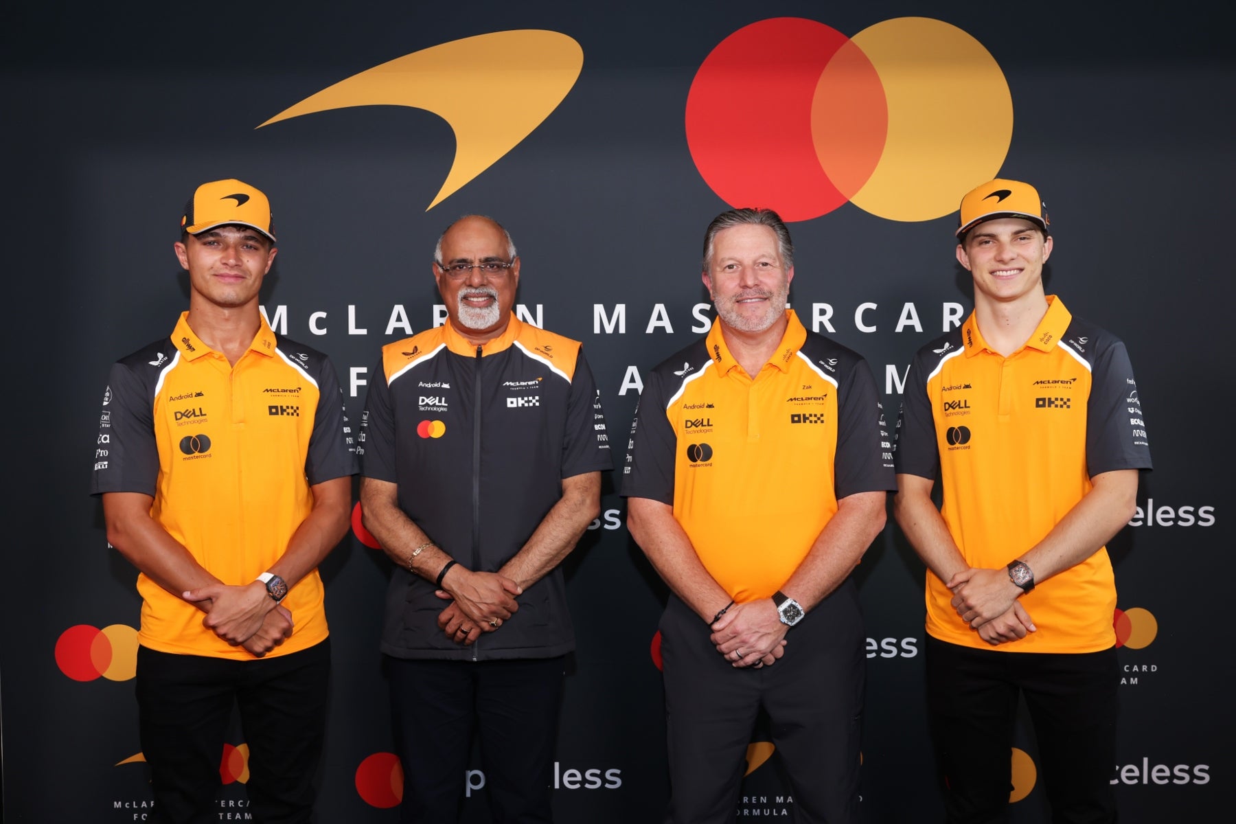 Four individuals in McLaren F1 team attire standing in front of a dark background with logos. - Endless Racegear